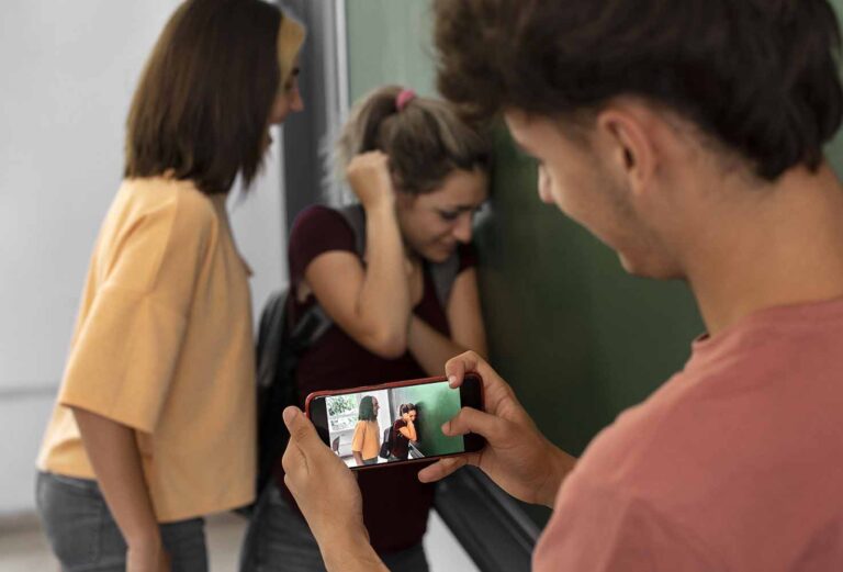 Person holding a smartphone and filming two girls in a hallway; one girl looks distressed with her head down and hand to her hair.