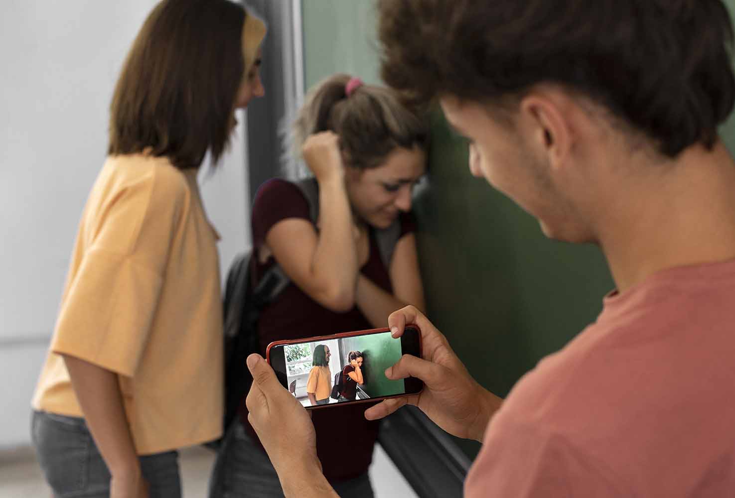 Person holding a smartphone and filming two girls in a hallway one girl looks distressed with her head down and hand to her hair