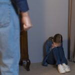 Young girl sitting on the floor with knees drawn to chest hands over her face looking distressed a blurred adult figure stands in the foreground