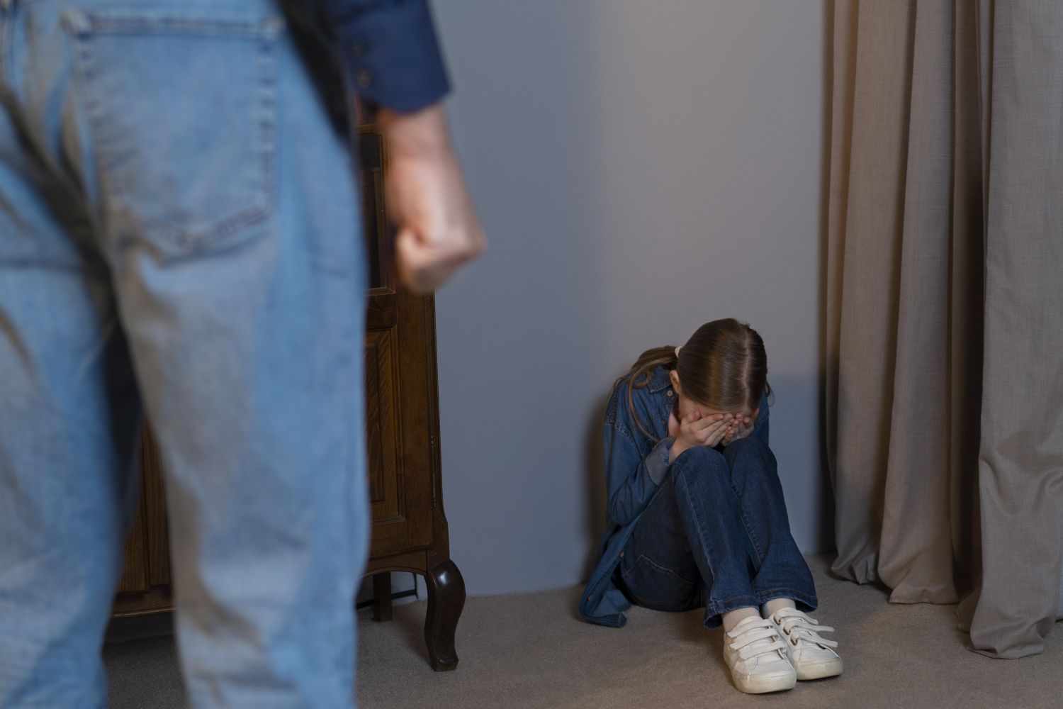 Young girl sitting on the floor with knees drawn to chest hands over her face looking distressed a blurred adult figure stands in the foreground