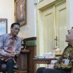 Two men in batik shirts sit and chat in a richly decorated room with gold trim and framed portraits