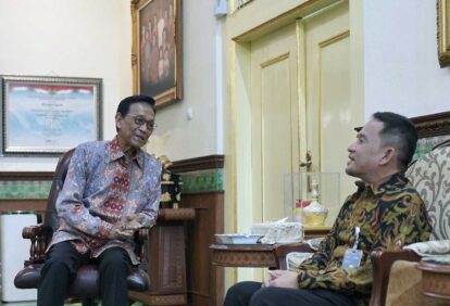 Two men in batik shirts sit and chat in a richly decorated room with gold trim and framed portraits
