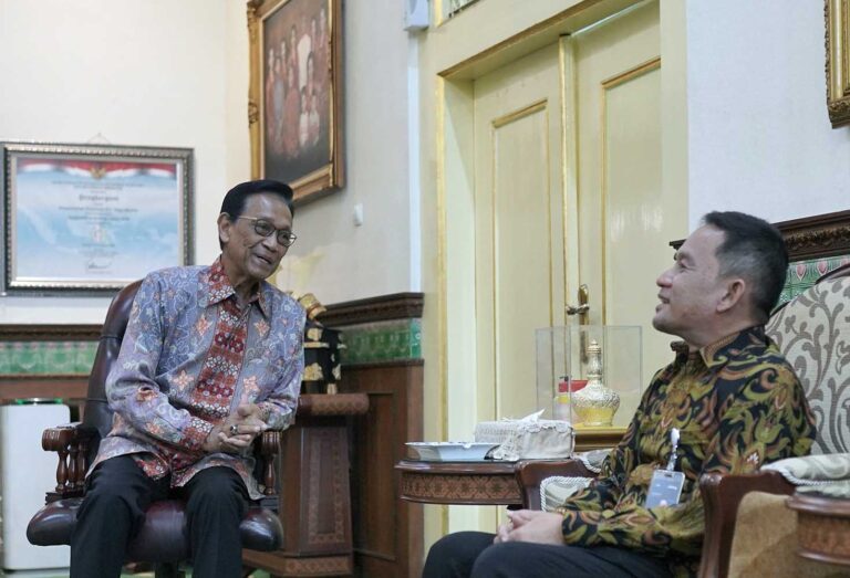 Two men in batik shirts sit and chat in a richly decorated room with gold trim and framed portraits.