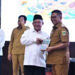 Group of men on stage at an awards ceremony central man in a white shirt and black cap shakes hands with a brown uniformed officer who holds a booklet
