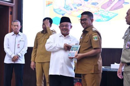 Group of men on stage at an awards ceremony; central man in a white shirt and black cap shakes hands with a brown-uniformed officer who holds a booklet.