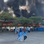 Group of people in blue uniforms evacuating as a large fire and thick black smoke engulfs a building behind them