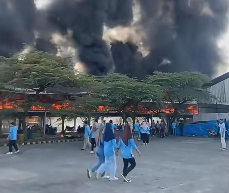 Group of people in blue uniforms evacuating as a large fire and thick black smoke engulfs a building behind them.