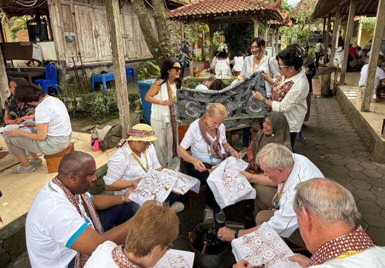 Group of people from different ages weaving intricate brown-on-white fabric panels in an outdoor courtyard workshop.
