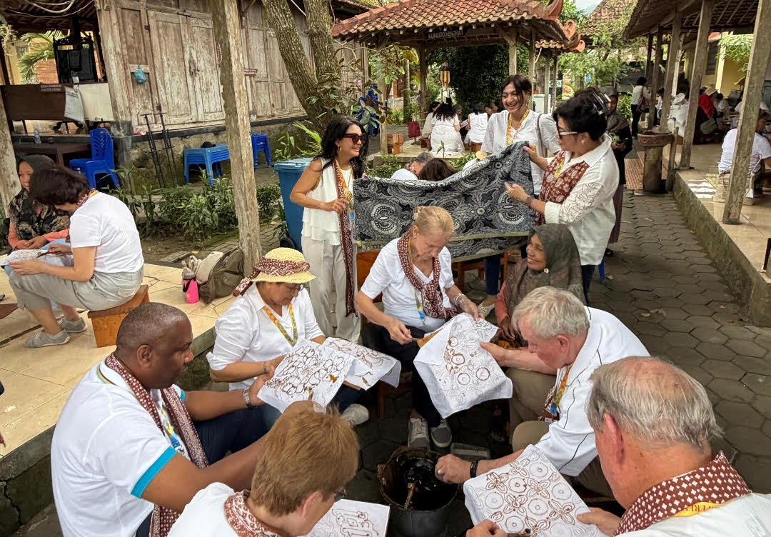 Group of people from different ages weaving intricate brown on white fabric panels in an outdoor courtyard workshop