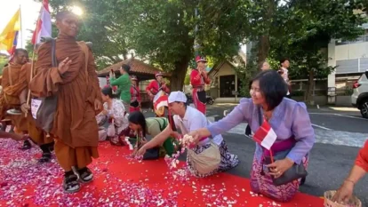 Monks in orange robes walking past a red carpet as people kneel and shower petals; Indonesian flags in the background.