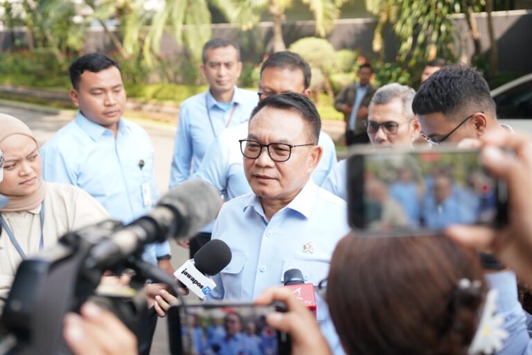 Man in a light-blue shirt being interviewed by the press outdoors, surrounded by reporters and cameras.