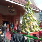 Crowd in traditional attire gathered around a tall decorative tree wrapped with hanging gift packets at a temple entrance