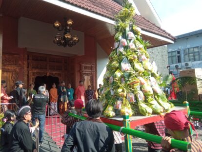 Crowd in traditional attire gathered around a tall decorative tree wrapped with hanging gift packets at a temple entrance.