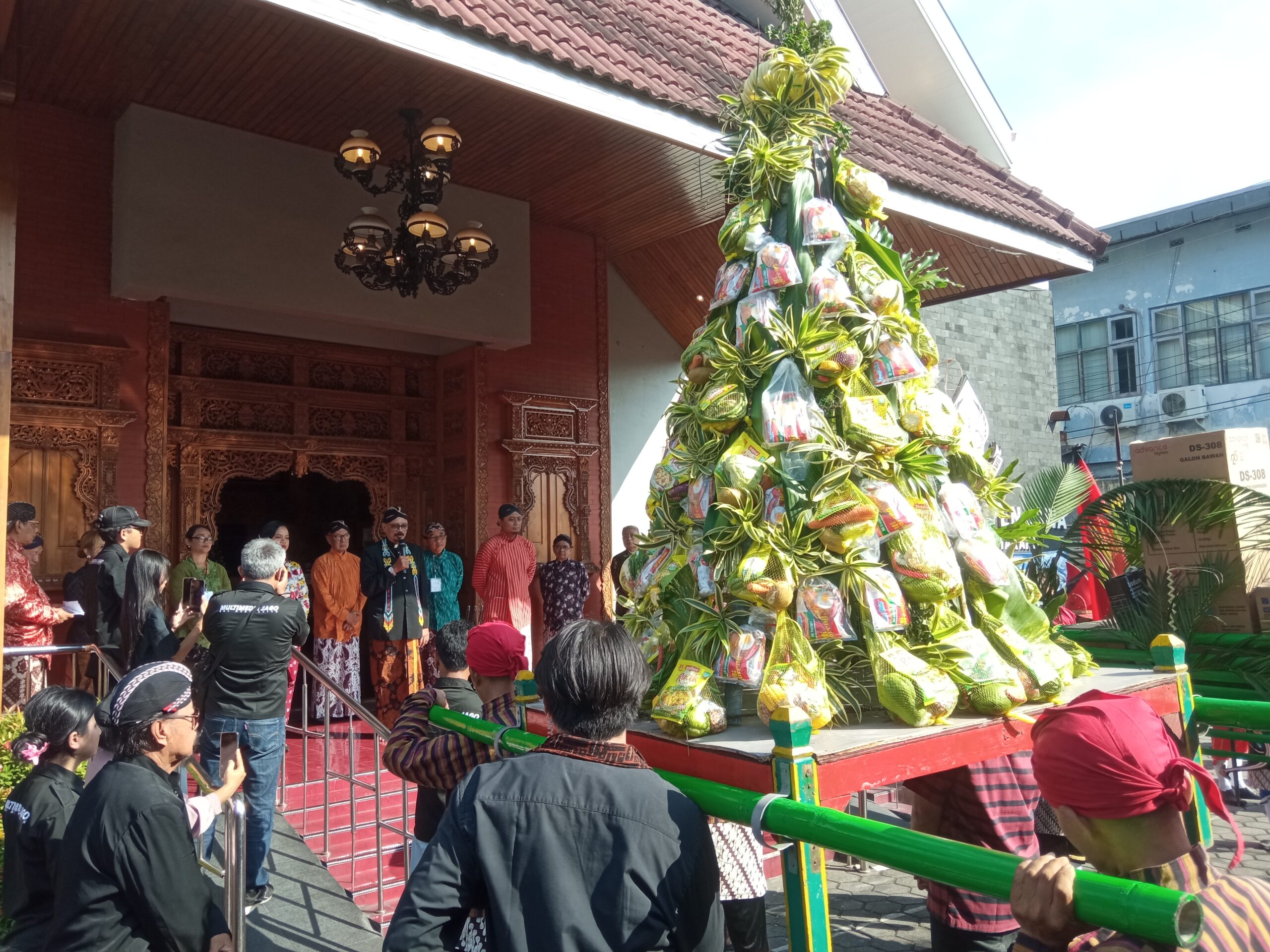 Gunungan catrawarta Crowd in traditional attire gathered around a tall decorative tree wrapped with hanging gift packets at a temple entrance