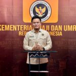 Male indonesian official in a beige uniform standing at a podium in front of a backdrop reading kementerian haji dan umrah republik indonesia with the ministry emblem above him
