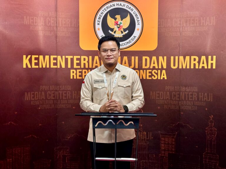 Male Indonesian official in a beige uniform standing at a podium, in front of a backdrop reading 'Kementerian Haji dan Umrah Republik Indonesia' with the ministry emblem above him