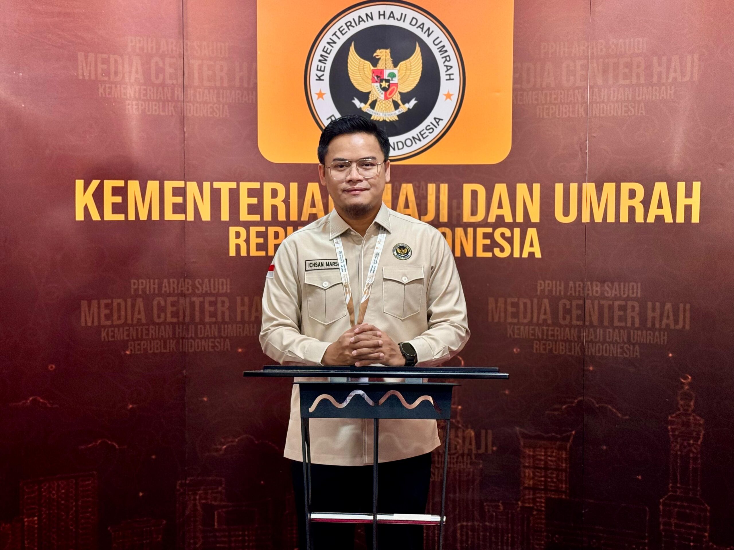 Haj catrawarta Male indonesian official in a beige uniform standing at a podium in front of a backdrop reading kementerian haji dan umrah republik indonesia with the ministry emblem above him