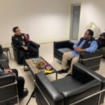 Four men in dark uniforms sit in a modern waiting room around a coffee table with water bottles and a flower arrangementthey appear to be talking