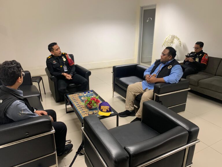 Four men in dark uniforms sit in a modern waiting room around a coffee table with water bottles and a flower arrangement—they appear to be talking.