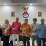 Eight people stand in a row in an office raising fists in a group pose with national emblems and portraits on the wall behind them