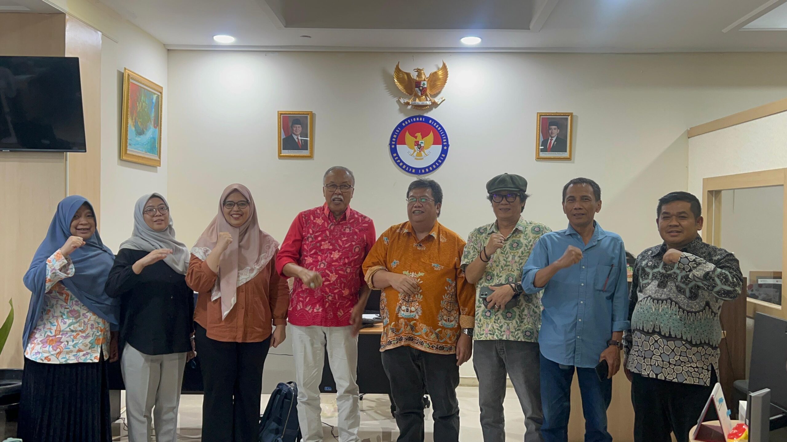 Eight people stand in a row in an office raising fists in a group pose with national emblems and portraits on the wall behind them