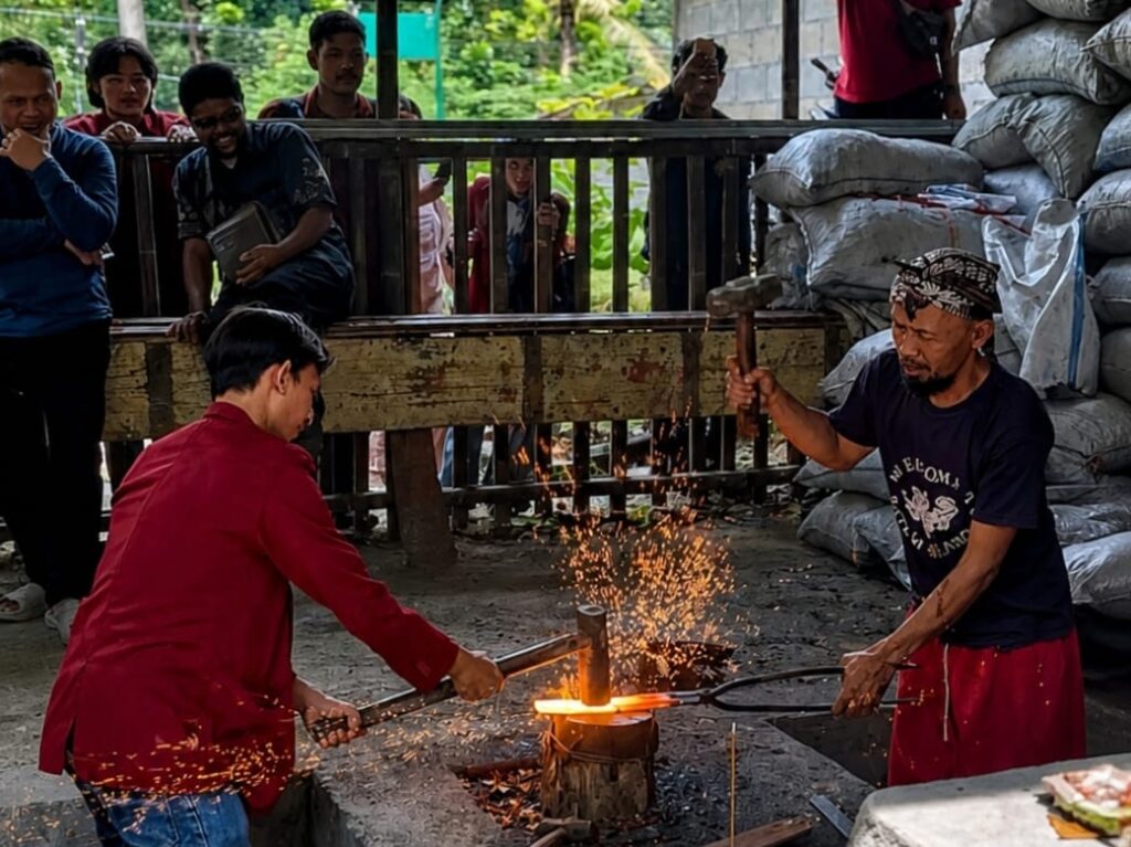 Two men hammering glowing metal on an anvil in an outdoor workshop while sparks fly as onlookers stand behind a wooden barrier and sacks lie nearby far in the background