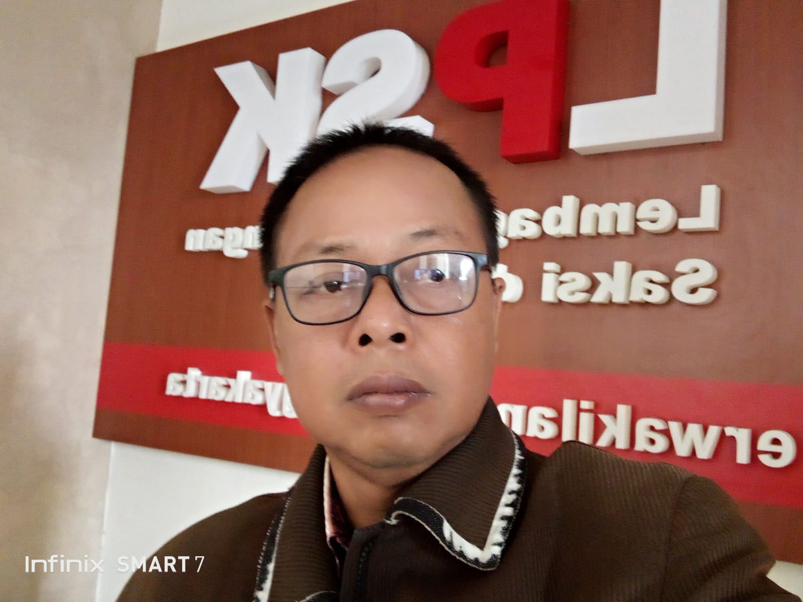 Img20260502wa0021 catrawarta Man wearing glasses takes a close up selfie in front of a brown wall with large white and red company sign behind him
