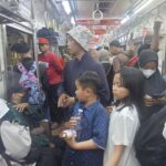 Crowded subway car with standing passengers holding straps several wear masks a boy in a blue polka dot shirt holds a bottle