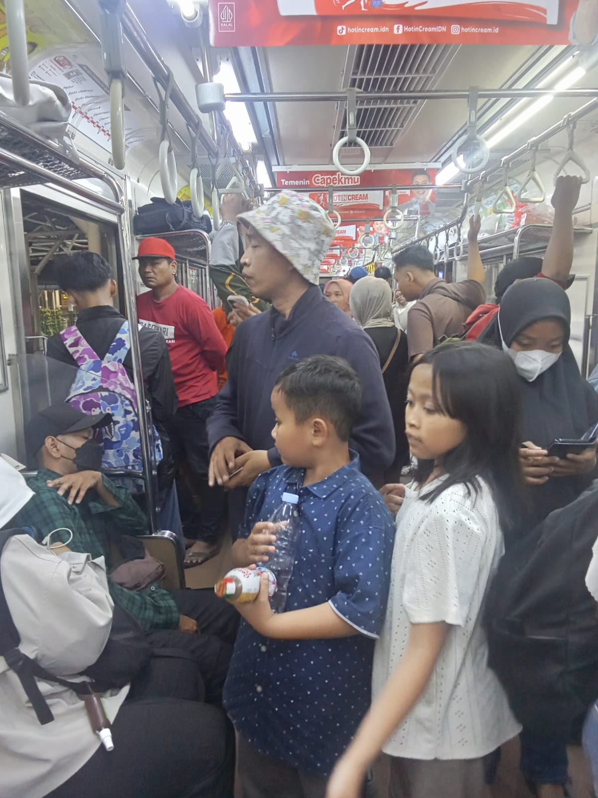 Crowded subway car with standing passengers holding straps several wear masks a boy in a blue polka dot shirt holds a bottle