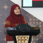 Woman in a red hijab and patterned red top speaks at a black podium with a gold decorative panel