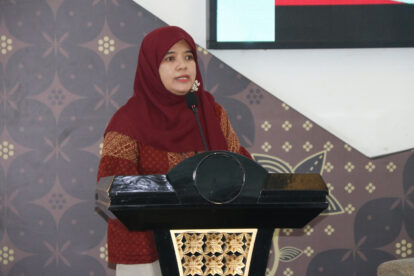 Woman in a red hijab and patterned red top speaks at a black podium with a gold decorative panel.