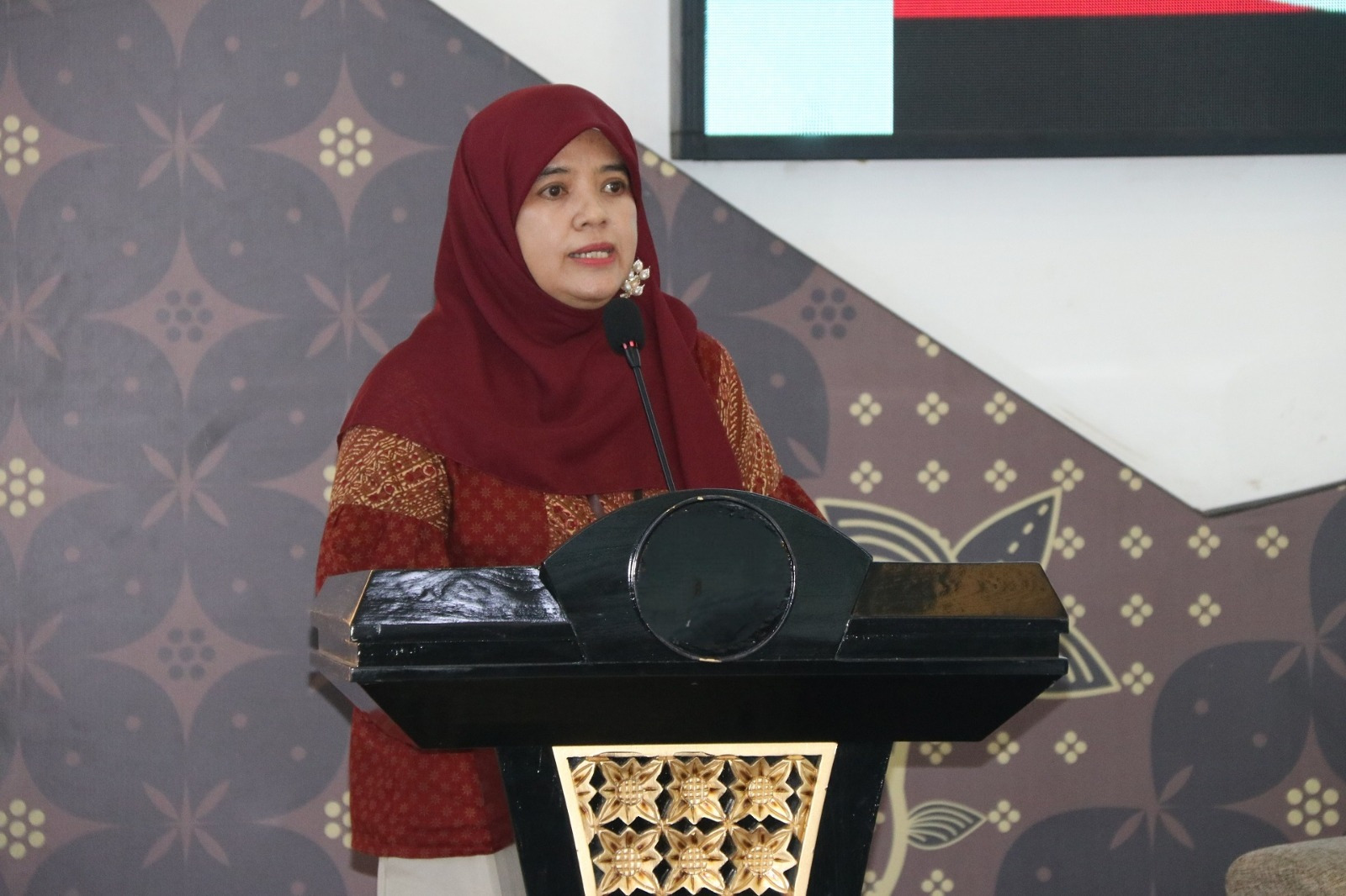 Woman in a red hijab and patterned red top speaks at a black podium with a gold decorative panel