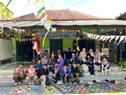 Group of people posing on steps outside a green building decorated with colorful banners for a community event, Indonesian flag in view.