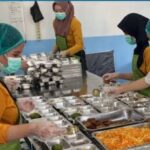 Cafeteria workers in hair nets and masks serve meals from a stainless steel serving line trays hold a variety of dishes
