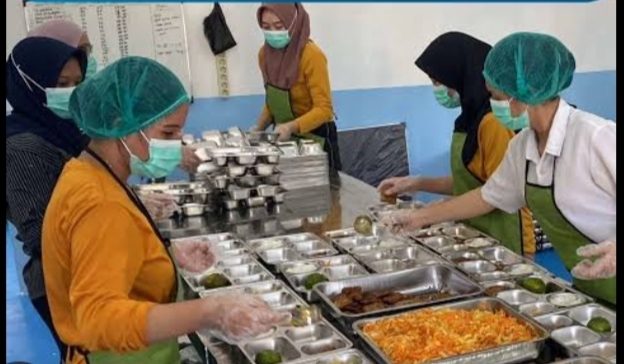 Cafeteria workers in hair nets and masks serve meals from a stainless steel serving line; trays hold a variety of dishes.