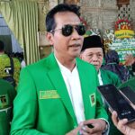 Man in a bright green suit being interviewed by reporters wearing sunglasses flanked by two men in green uniforms at a ceremony