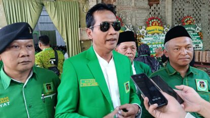 Man in a bright green suit being interviewed by reporters, wearing sunglasses, flanked by two men in green uniforms at a ceremony.
