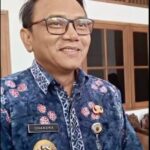 Smiling man in a blue batik uniform with a name badge chandra and several gold insignia standing indoors near wooden windows