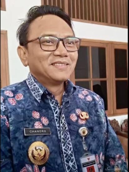 Smiling man in a blue batik uniform with a name badge 'Chandra' and several gold insignia, standing indoors near wooden windows.