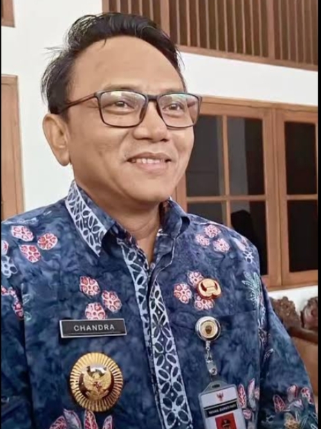 Img20260504wa0014 catrawarta Smiling man in a blue batik uniform with a name badge chandra and several gold insignia standing indoors near wooden windows