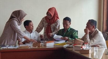Five colleagues sit around a cluttered conference table, discussing documents as two women in hijabs stand nearby.