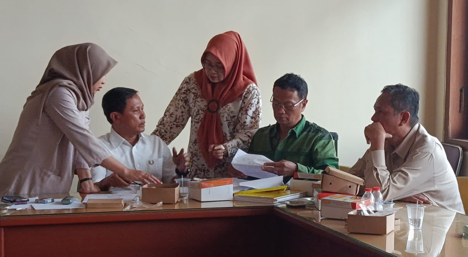 Five colleagues sit around a cluttered conference table discussing documents as two women in hijabs stand nearby