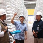 Three men wearing hard hats stand in a warehouse aisle between tall stacks of white sacks one gestures while another holds a blue bin