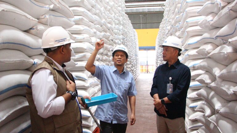 Three men wearing hard hats stand in a warehouse aisle between tall stacks of white sacks; one gestures while another holds a blue bin.