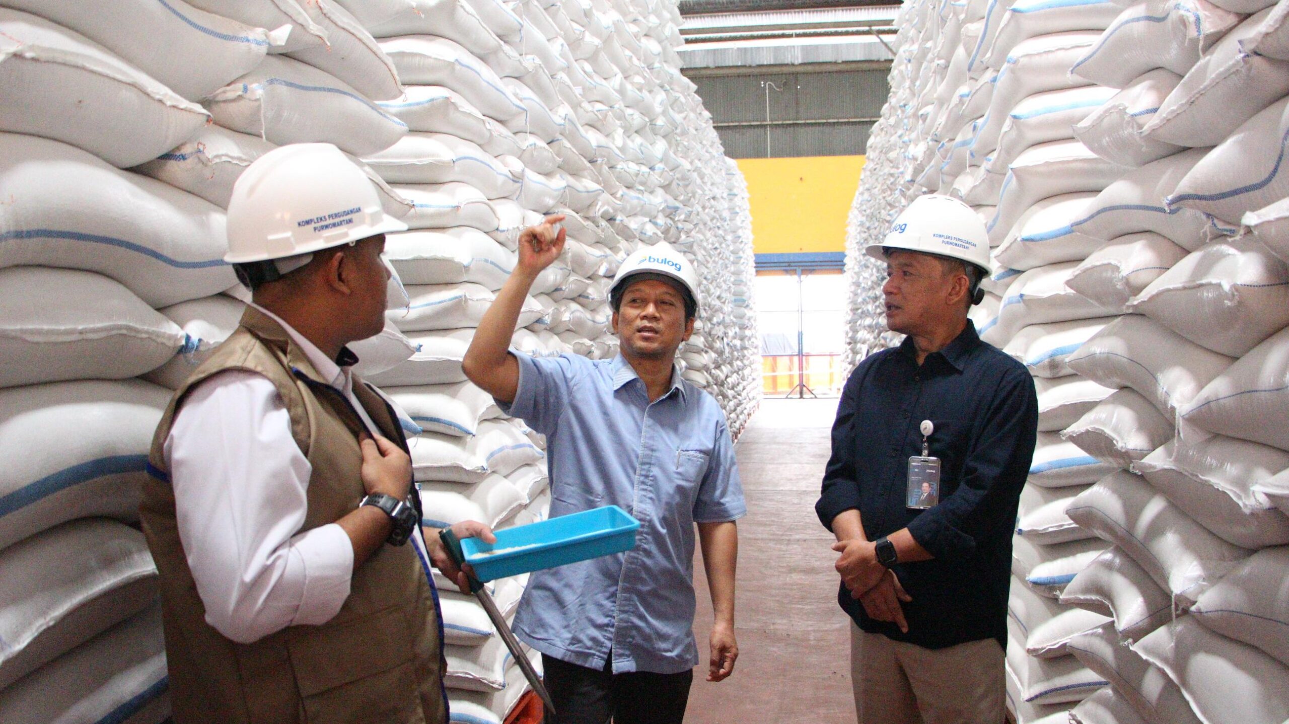 Three men wearing hard hats stand in a warehouse aisle between tall stacks of white sacks one gestures while another holds a blue bin