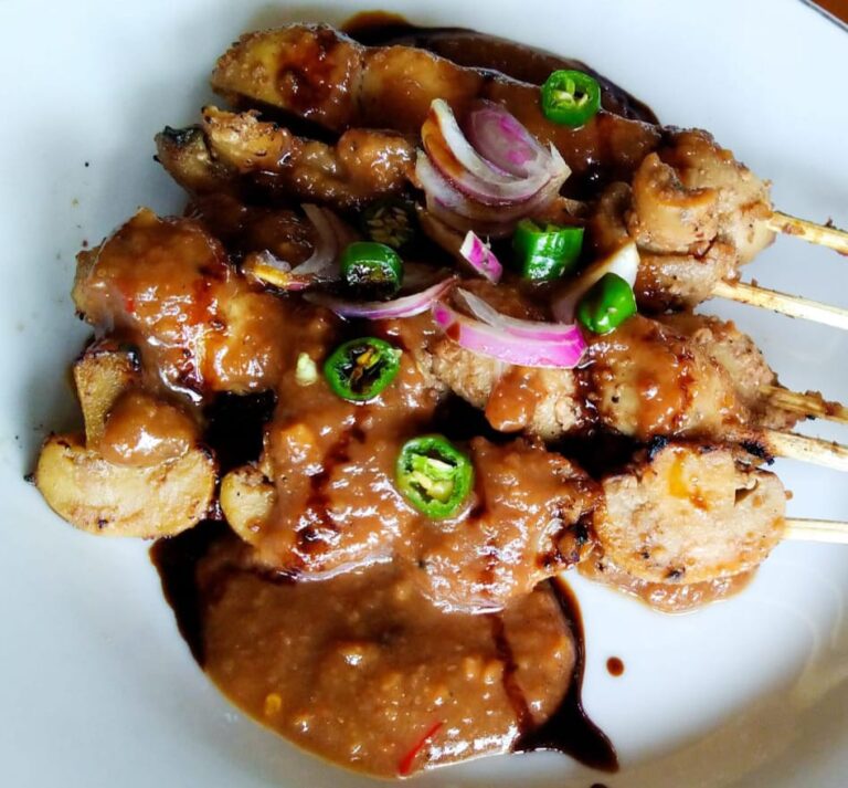 Skewered grilled meat with a glossy brown sauce, garnished with sliced red onions and jalapeño rings on a white plate.