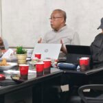 Three men sit around a cluttered meeting table with laptops snacks and drinks one gesturing while speaking