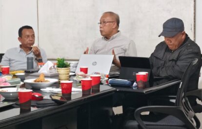 Three men sit around a cluttered meeting table with laptops snacks and drinks one gesturing while speaking