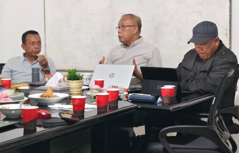 Three men sit around a cluttered meeting table with laptops, snacks, and drinks, one gesturing while speaking.