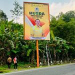 Large yellow campaign billboard on a wooden post near a road featuring a man in a yellow shirt and bold black text for musda dpd golkar surrounded by green plants and a ladder nearby